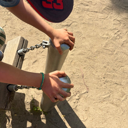 Kind trägt das Kids' Bracenet Kieler Bucht aus einem Strang türkisen Fischernetz und einem Strang buntem Fischernetz, größenverstellbar mit Magnetverschluss in Silber am Handgelenk und ist auf einem Spielplatz. 