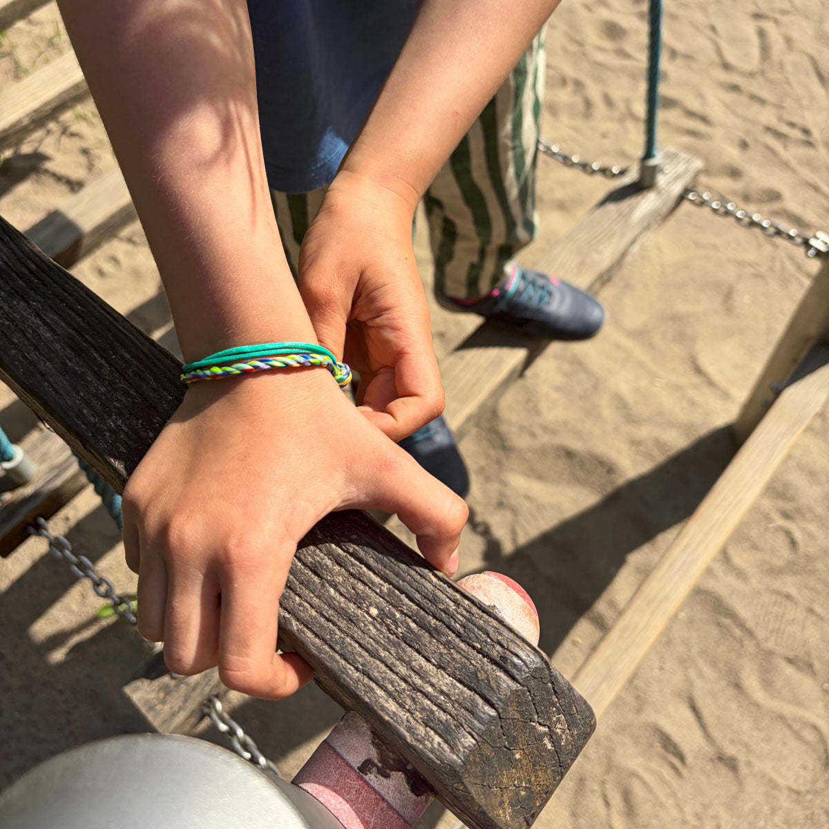 Kind trägt das Kids' Bracenet Kieler Bucht aus einem Strang türkisen Fischernetz und einem Strang buntem Fischernetz, größenverstellbar mit Magnetverschluss in Silber Am Handgelenk und hält sich an einem Holzpfosten fest. 