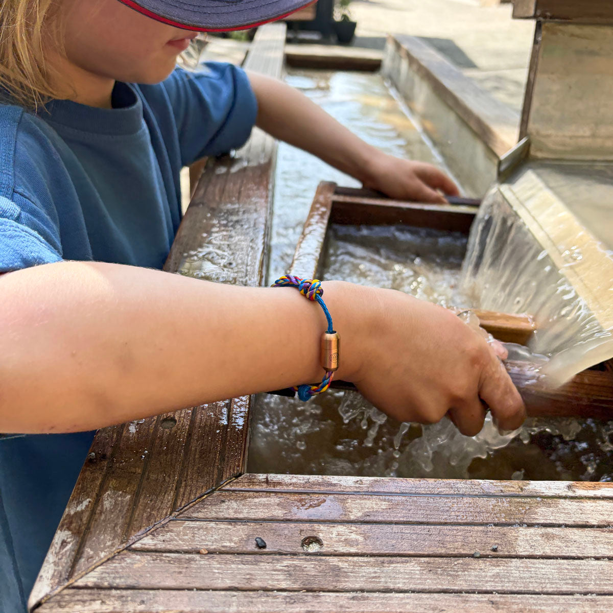 Kind trägt das Kids' Bracenet Lübecker Bucht aus einem Strang blauem Fischernetz und einem Strang buntem Fischernetz, größenverstellbar mit Magnetverschluss in Roségold am Handgelenk und spielt mit Wasser. 