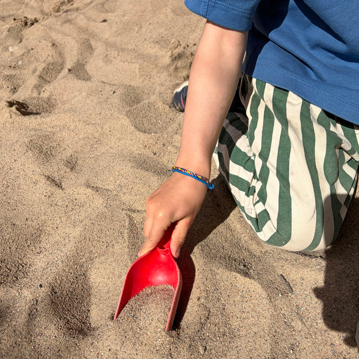 Kind trägt das Kids' Bracenet Lübecker Bucht aus einem Strang blauem Fischernetz und einem Strang buntem Fischernetz, größenverstellbar mit Magnetverschluss in Roségold am Handgelenk und spielt im Sand. 
