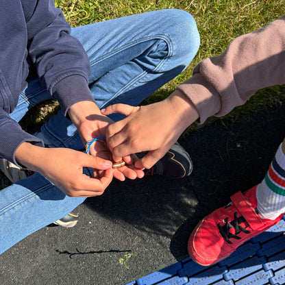 Mutter hält das Kids' Bracenet Lübecker Bucht aus einem Strang blauem Fischernetz und einem Strang buntem Fischernetz, größenverstellbar mit Magnetverschluss in Roségold in der Hand und Kind greift danach. 