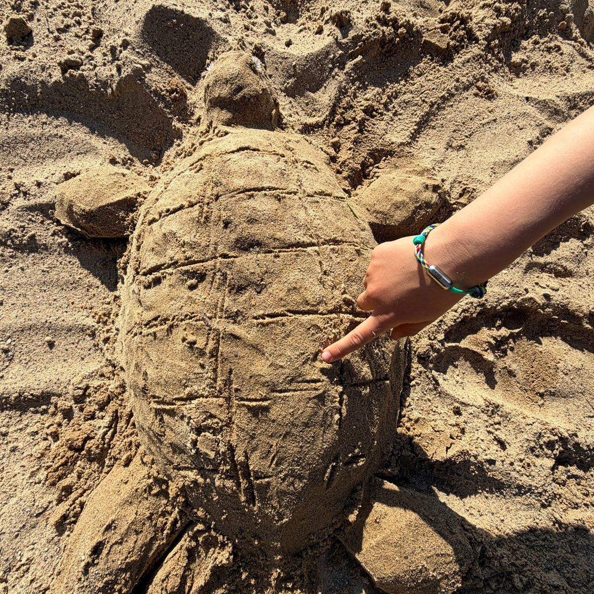 Kind trägt das Kids' Bracenet Kieler Bucht aus einem Strang türkisen Fischernetz und einem Strang buntem Fischernetz, größenverstellbar mit Magnetverschluss in Silber am Handgelenk und baut eine Schildkröte in den Sand. 