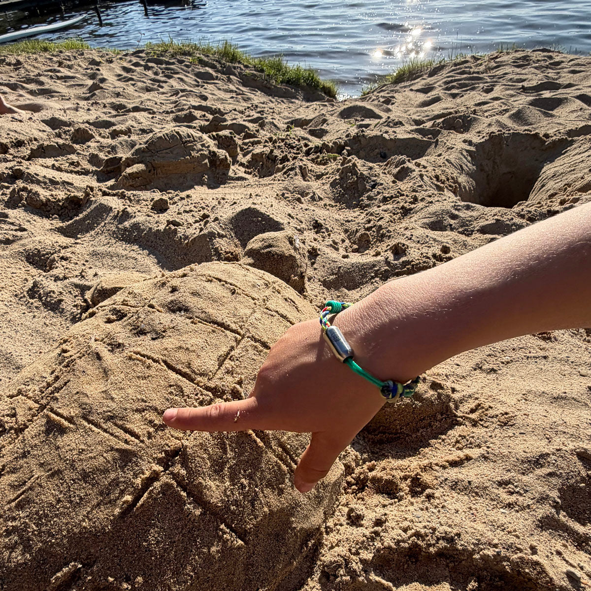 Kind trägt das Kids' Bracenet Kieler Bucht aus einem Strang türkisen Fischernetz und einem Strang buntem Fischernetz, größenverstellbar mit Magnetverschluss in Silber am Handgelenk und spielt im Sand. 
