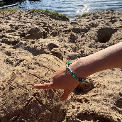 Kind trägt das Kids' Bracenet Kieler Bucht aus einem Strang türkisen Fischernetz und einem Strang buntem Fischernetz, größenverstellbar mit Magnetverschluss in Silber am Handgelenk und spielt im Sand. 