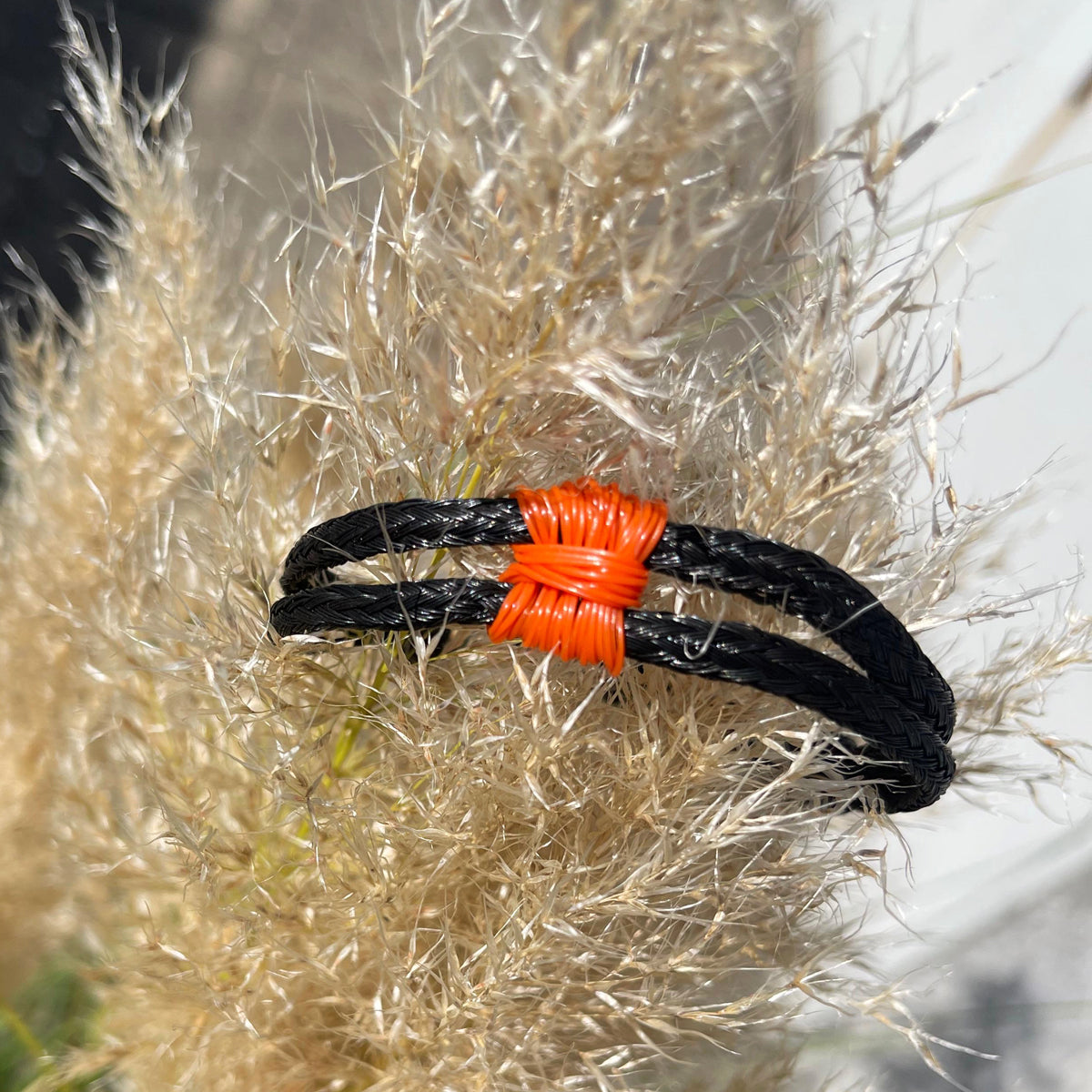 Das BRACENET Upcycling Armband Pumpkin Bay II aus schwarzen alten Fischernetzen mit einem Knoten aus orangenen Dolly Ropes und einem schwarzen Magnetverschluss ist auf eine Pflanze gesteckt. Man sieht den orangenen Knoten. 