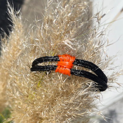 Das BRACENET Upcycling Armband Pumpkin Bay II aus schwarzen alten Fischernetzen mit einem Knoten aus orangenen Dolly Ropes und einem schwarzen Magnetverschluss ist auf eine Pflanze gesteckt. Man sieht den orangenen Knoten. 