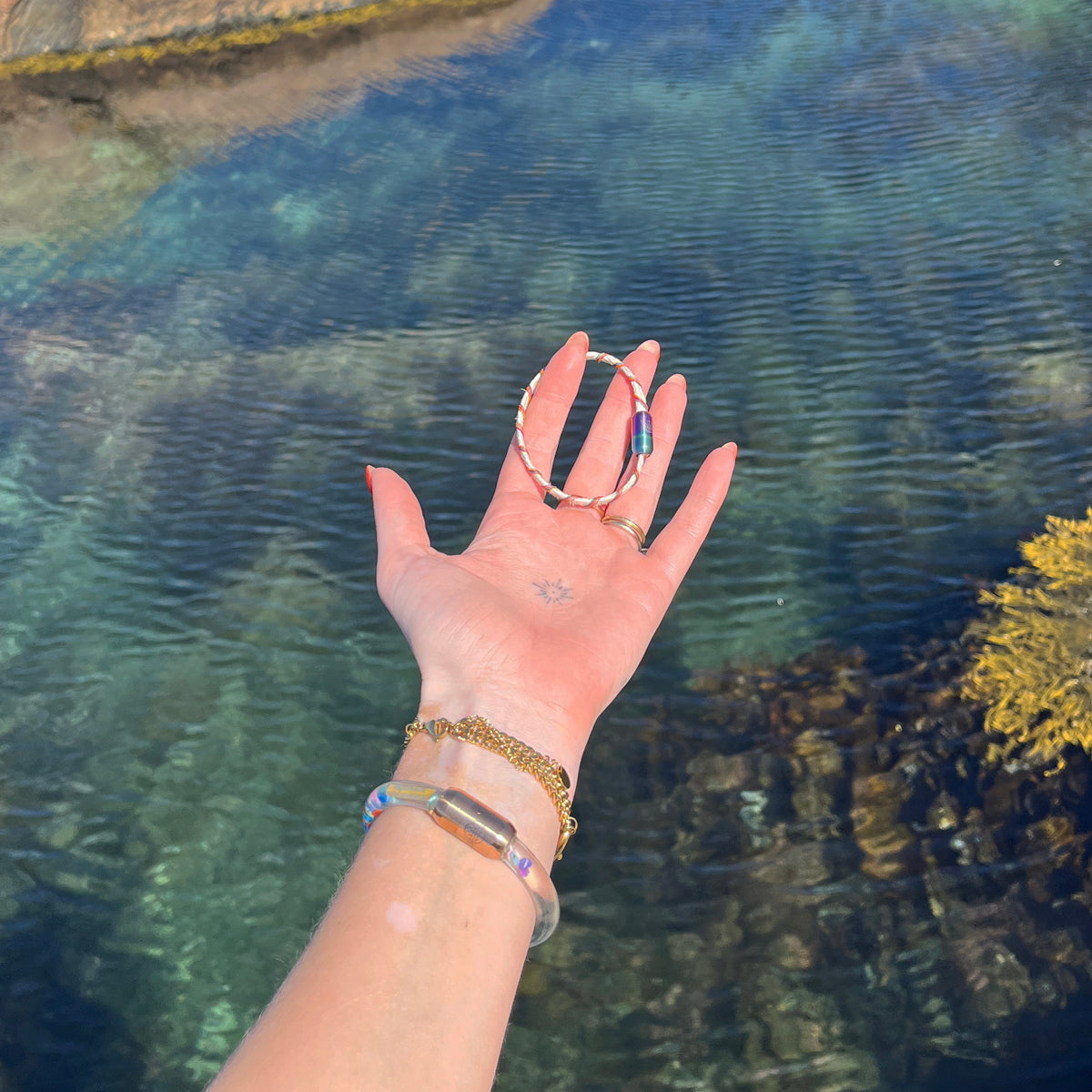 Person hält das BRACENET Upcycling Armband Rainbow Bay aus alten weißen Fischernetzen mit bunten Dolly Ropes umwickelt und bunten Magnetverschluss mit Farbverlauf in der Hand, im Hintergrund klares Fjordwasser