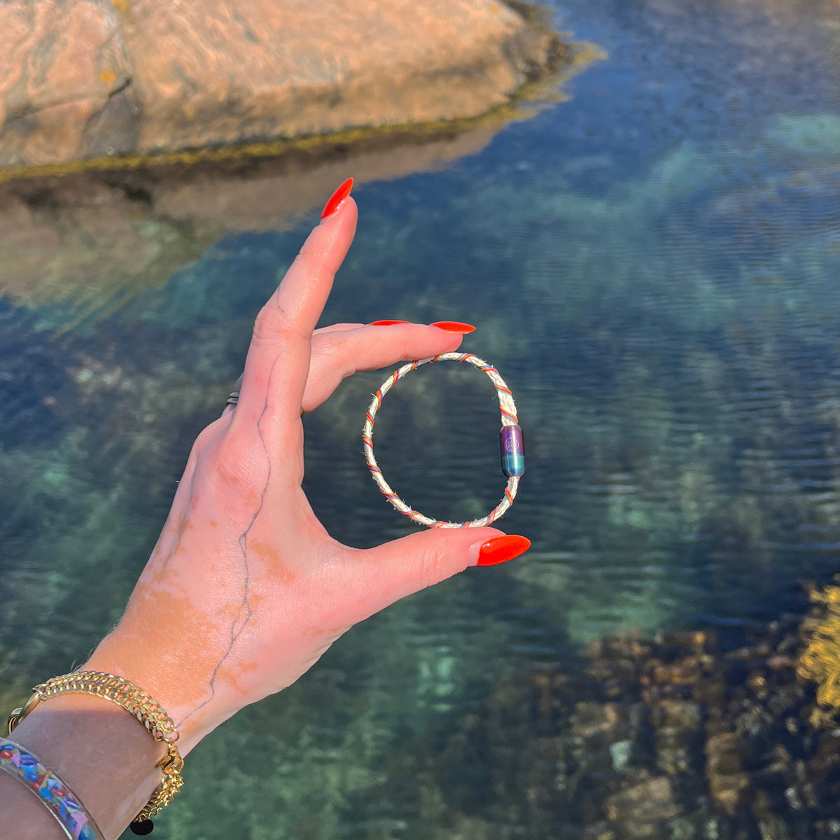 Person hält das BRACENET Upcycling Armband Rainbow Bay aus alten weißen Fischernetzen mit bunten Dolly Ropes umwickelt und bunten Magnetverschluss mit Farbverlauf in den Händen, im Hintergrund ein Fjord mit glasklarem Wasser
