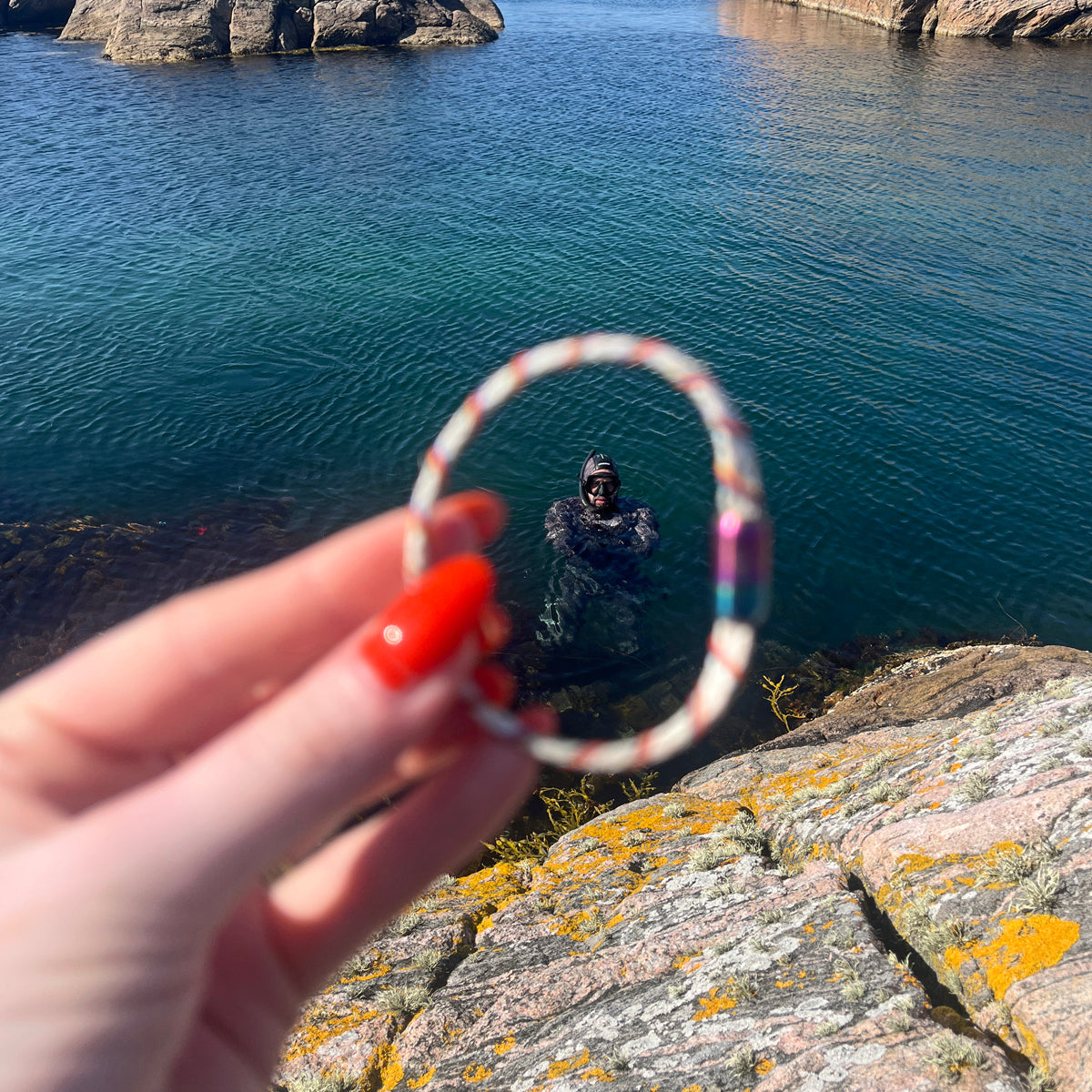 Person hält das BRACENET Upcycling Armband Rainbow Bay aus alten weißen Fischernetzen mit bunten Dolly Ropes umwickelt und bunten Magnetverschluss mit Farbverlauf in den Vordergrund, Fokus durch das Armband liegt auf den Taucher im Wasser