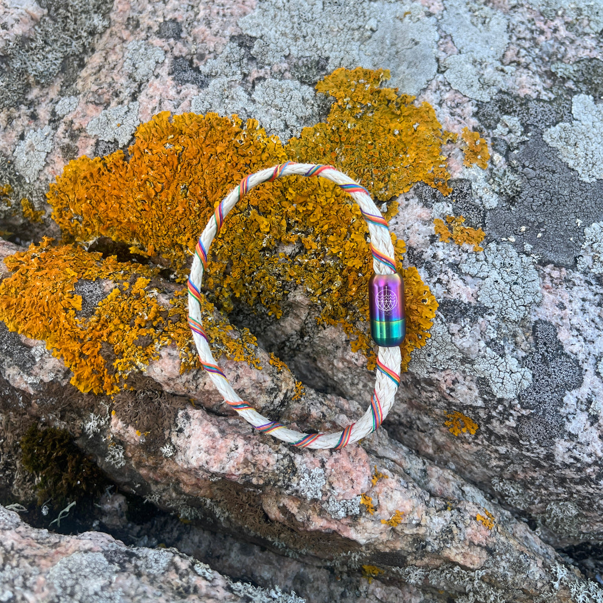 Das BRACENET Upcycling Armband Rainbow Bay aus alten weißen Fischernetzen mit bunten Dolly Ropes umwickelt und bunten Magnetverschluss mit Farbverlauf liegt auf einem Stein