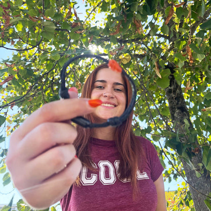 Person hält das BRACENET Upcycling Armband Pumpkin Bay II aus schwarzen alten Fischernetzen mit einem Knoten aus orangenen Dolly Ropes und einem schwarzen Magnetverschluss vor das Gesicht und lacht. Im Hintergrund sieht man einen Baum. 