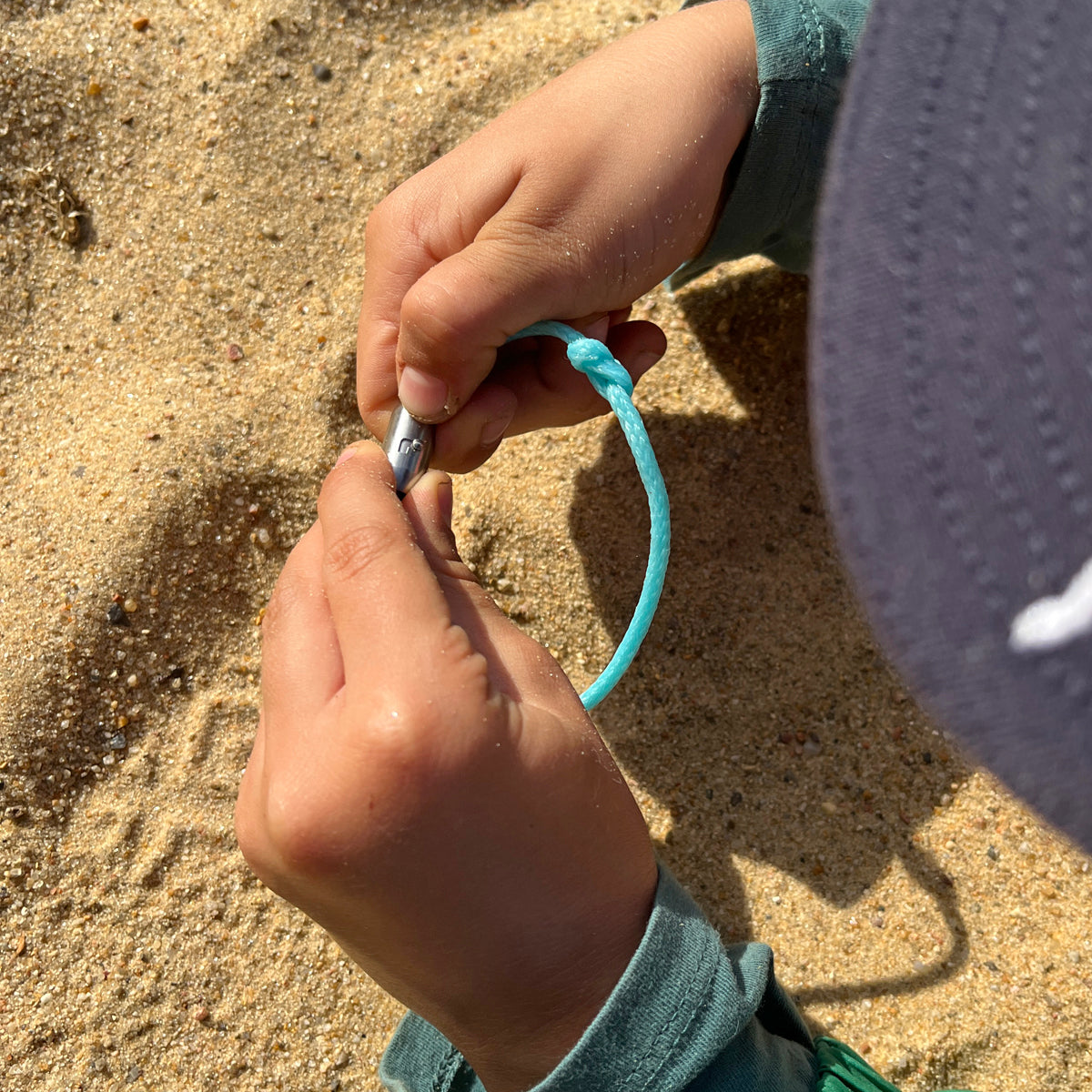 Kind öffnet das BRACENET Upcycling Armband Glacier Bay aus eisblauen Fischernetzen mit silbernen Bajonettverschluss, im Hintergrund Sand