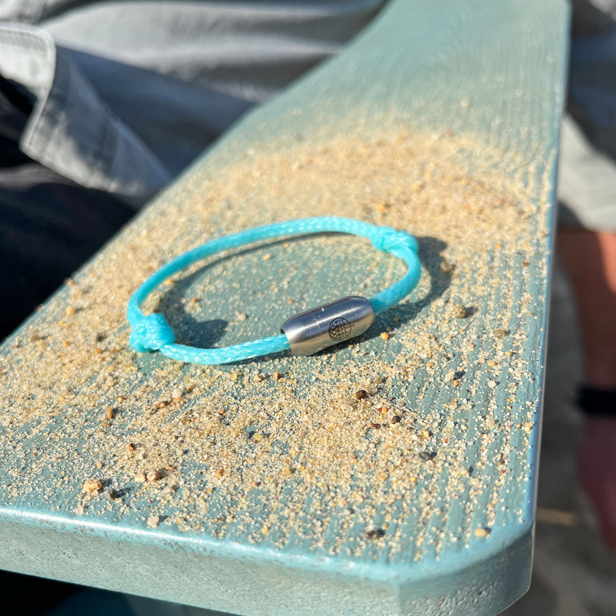 Das BRACENET Upcycling Armband Glacier Bay aus eisblauen Fischernetzen und 2 Knoten mit silbernen Bajonettverschluss liegt auf einem Holzbrett mit Sand