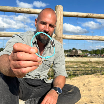 Person hält das BRACENET Upcycling Armband Glacier Bay aus eisblauen Fischernetzen und 2 Knoten mit silbernen Bajonettverschluss in die Kamera, im Hintergrund Sand