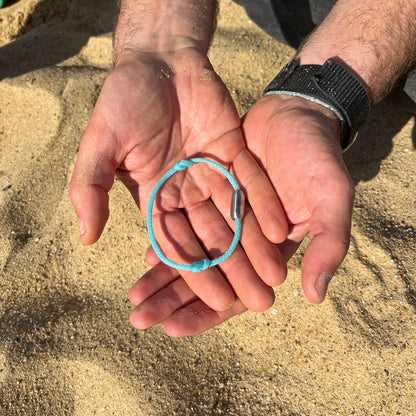 Person hält das BRACENET Upcycling Armband Glacier Bay aus eisblauen Fischernetzen  mit silbernen Bajonettverschluss in den Händen, im Hintergrund Sand