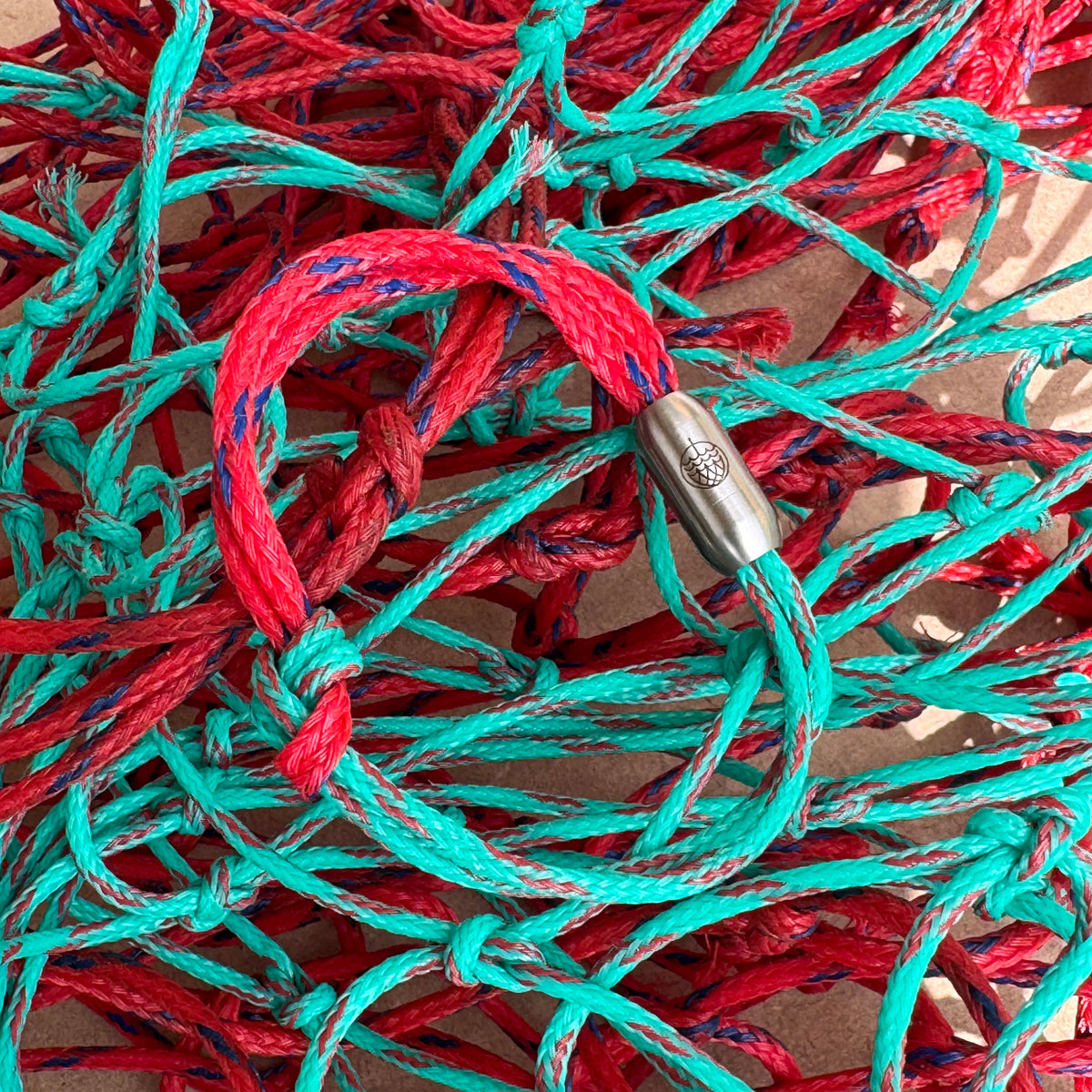 April Bracenet Gulf of Mannar aus Fischernetz in Rot und Türkis mit Knoten und mit Bajonettverschluss in Silber liegt auf Fischernetzen. 