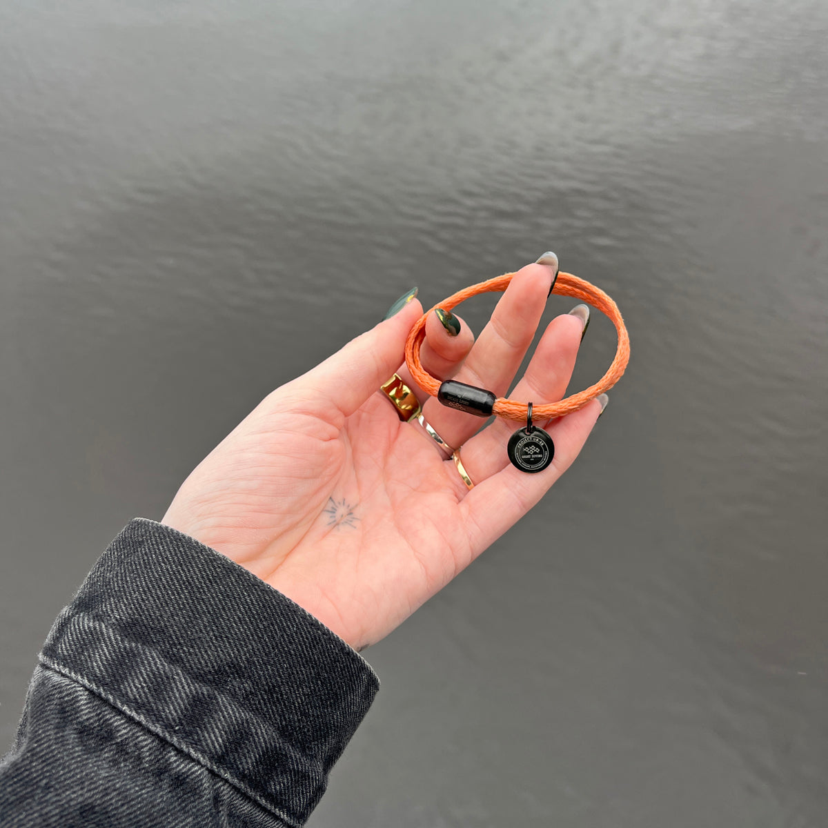 Person hält das BRACENET Upcycling Armband Ghost Diving USA aus orangenen alten Fischernetzen und einem schwarzen Magnetverschluss und Edelstahlanhänger in den Händen, im Hintergrund Wasser