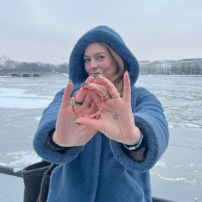 Person hält Februar Bracenet Amur Bay aus lachsfarbenen Fischernetz mit roten Dolly Ropes verziert und mit roségoldenen Magnetverschluss in beiden Händen vor sich. 
