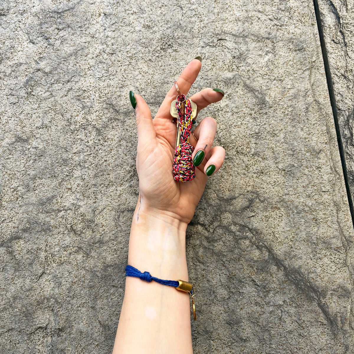 Perosn hält den BRACENET Schlüsselanhänger Gulf of California aus bunten Fischereiseilen mit Makramee und mit roségoldenem Schlüsselanhänger-Ring und Anhänger mit Delfin Gravur in der Hand und hat die Hand an der Wand. 
