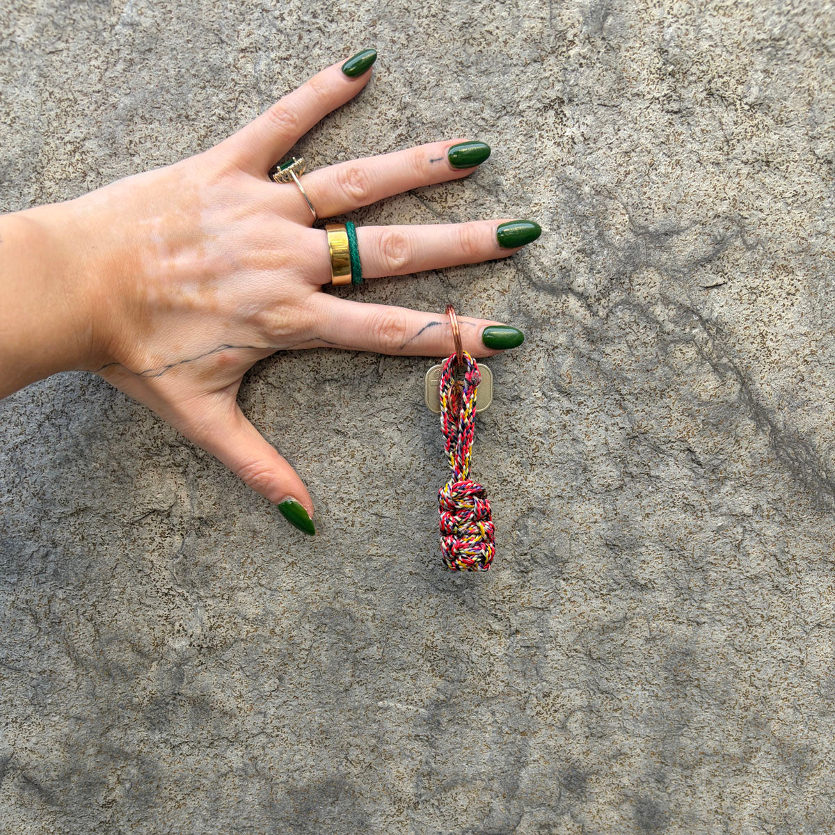 Person hat den BRACENET Schlüsselanhänger Gulf of California aus bunten Fischereiseilen mit Makramee und mit roségoldenem Schlüsselanhänger-Ring und Anhänger mit Delfin Gravur am Finger und hält die Hand vor eine Wand. 