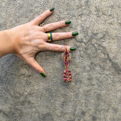 Person hat den BRACENET Schlüsselanhänger Gulf of California aus bunten Fischereiseilen mit Makramee und mit roségoldenem Schlüsselanhänger-Ring und Anhänger mit Delfin Gravur am Finger und hält die Hand vor eine Wand. 