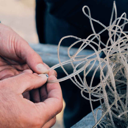Person befestigt das Alles für die Tiere immer - Robbe "Walvis" Armband aus transparenten Langleinen von BRACENET mit Drehverschluss in silber und "Alles für die Tiere, immer" Gravur mit Robben-Motiv an einem Stück unverarbeitetem Netz. 