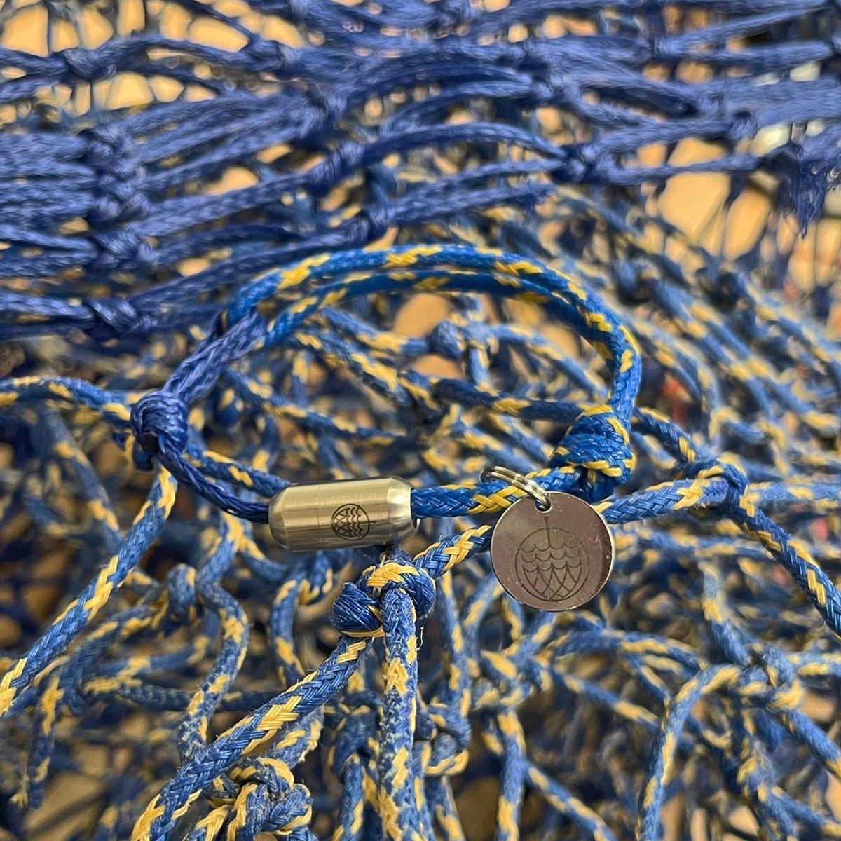 Das BRACENET Upcycling Armband Insel Sylt II aus alten Fischernetzen in dunkelblau und blau-gelb, mit einem silbernen Magnetverschluss und Anhänger mit einer Inselgravur von Sylt liegt auf den Fischernetzen aus denen es gefertigt wurde. 