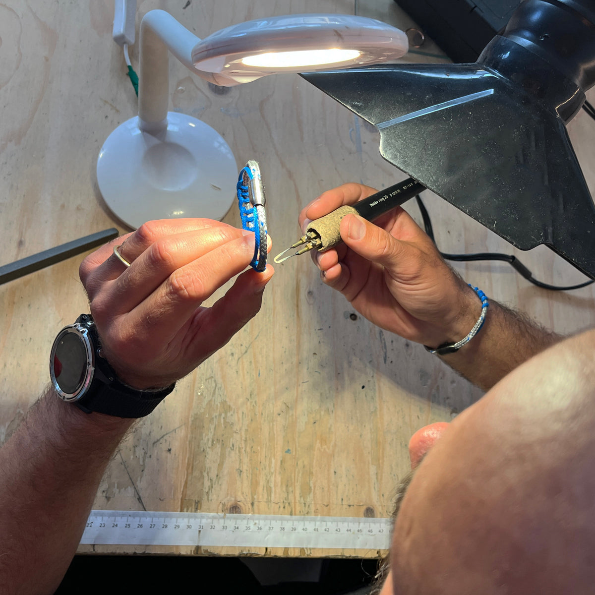 Das BRACENET Upcycling Armband Scotia Sea aus grauen und blauen alten Fischernetzen wird gefertigt. Die Person die das Armband mit dem Lötkolben fertigt trägt am Handgelenk ebenfalls ein Scotia Sea Bracenet mit schwarzem Bajonettverschluss. 