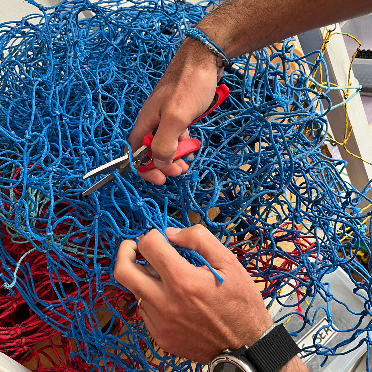 Person BRACENET Upcycling Armband Scotia Sea aus grauen und blauen alten Fischernetzen am Handgelenk und schneidet ein Stück blaues Netz. 