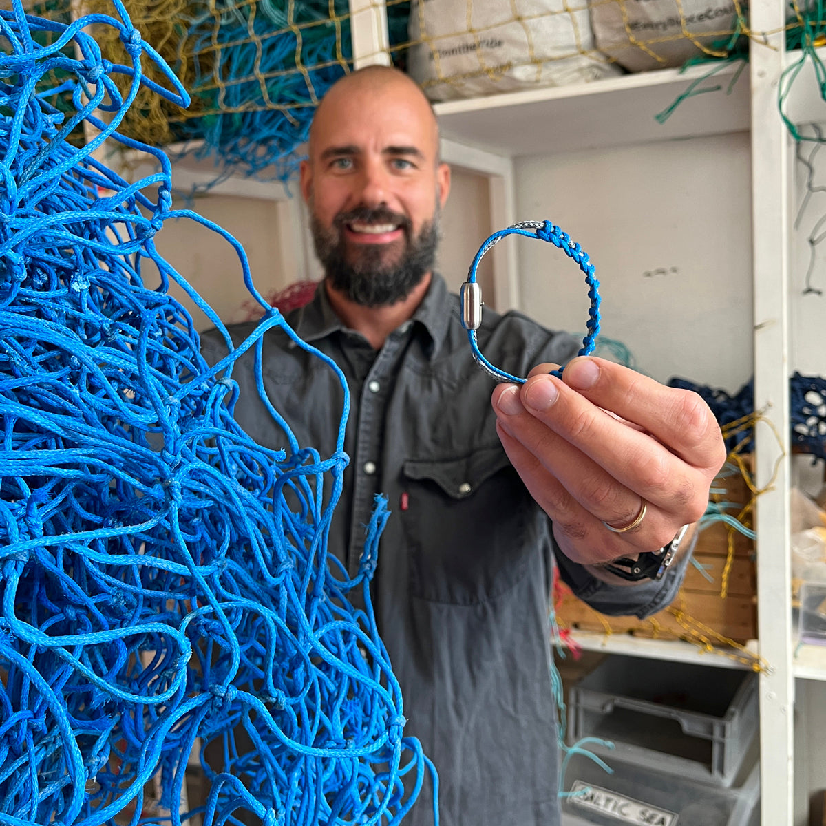 Person hält BRACENET Upcycling Armband Scotia Sea aus grauen und blauen alten Fischernetzen mit einem silbernen Bajonettverschluss vor sich in der Hand. In der anderen Hand hält die Person das blaue Netz. Die Person lacht.