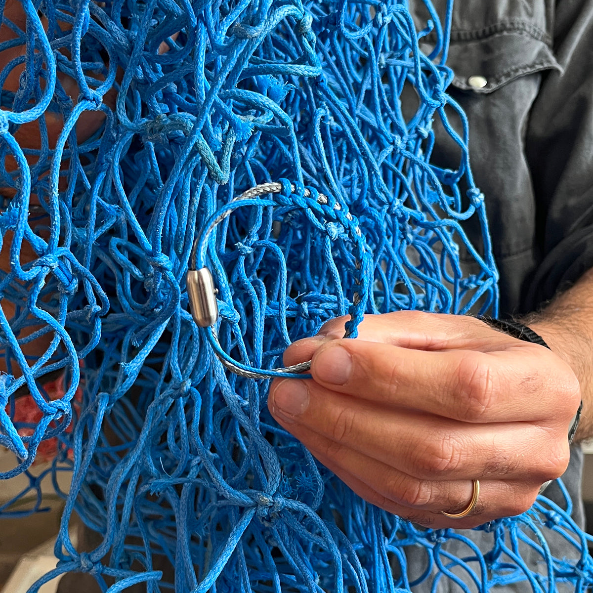 Person hält das BRACENET Upcycling Armband Scotia Sea aus grauen und blauen alten Fischernetzen mit silbernen Bajonettverschluss vor das blaue Fischernetz.
