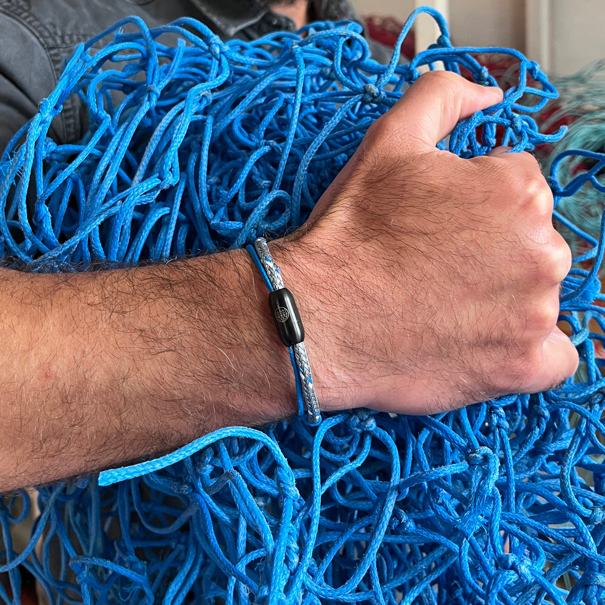Person trägt BRACENET Upcycling Armband Scotia Sea aus grauen und blauen alten Fischernetzen mit einem schwarzen Bajonettverschluss am Handgelenk und hält das blaue Fischernetz im Arm. 