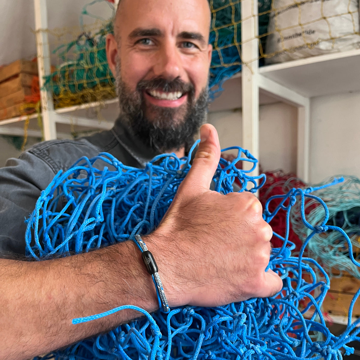 Person trägt BRACENET Upcycling Armband Scotia Sea aus grauen und blauen alten Fischernetzen mit einem schwarzen Bajonettverschluss am Handgelenk und zeigt einen Daumen nach oben. Im Arm hat die Person das blaue Netz. Die Person lacht. 