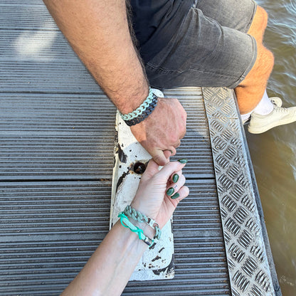 Person trägt das Juli Bracenet Zakynthos Navagio Bay aus Fischernetz in hellem Türkis mit Umwicklungen über Kreuz aus Fischernetz in dunklem Türkis und Magnetverschluss in Silber am Handgelenk und hat einen Finger mit der Hand einer anderen Person verhakt. 