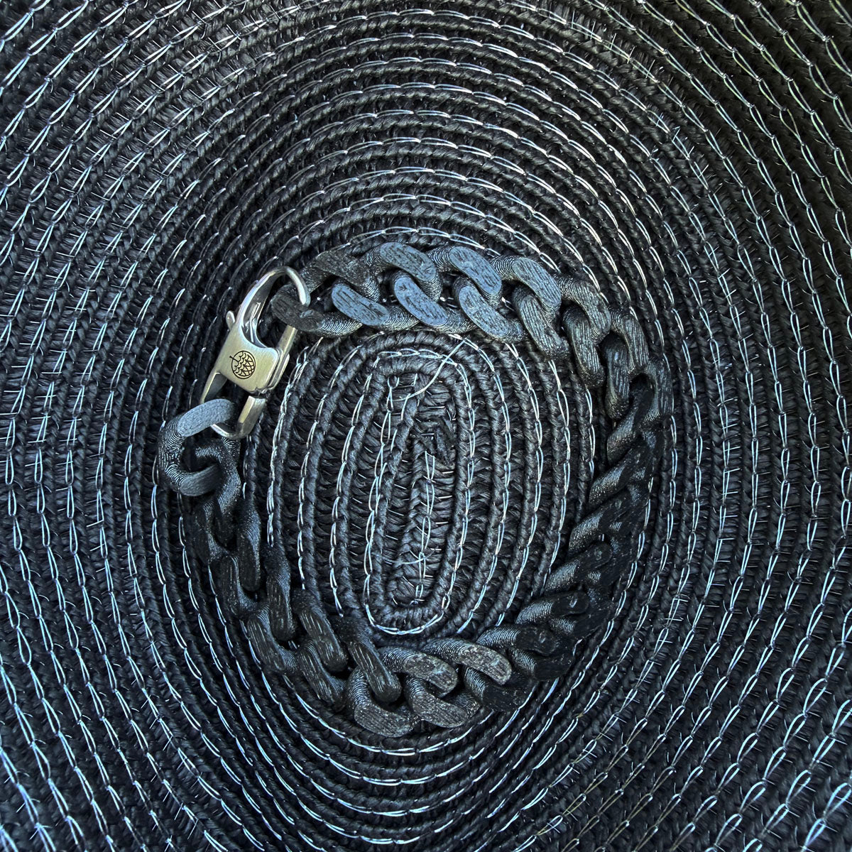Das SeaChain Bracenet in 3D Druck aus alten Fischernetzen hergestellt in Schwarz mit kleinem Clip Karabiner in Silber liegt auf einem Sonnenhut.