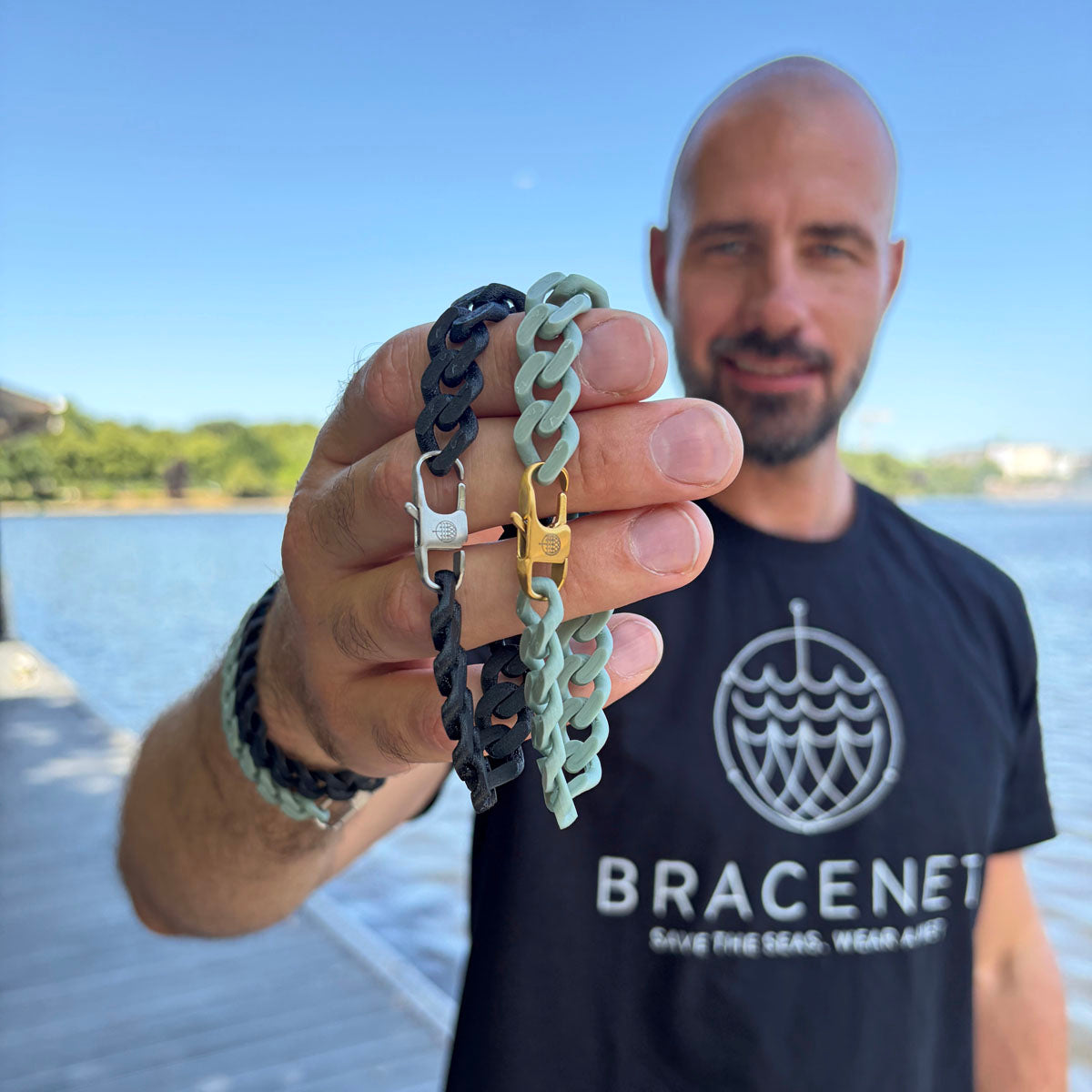 Person hält ein SeaChain Bracenet in 3D Druck aus alten Fischernetzen hergestellt in Blassgrün mit kleinem Clip Karabiner in Gold und ein SeaChain Bracenet in 3D Druck aus alten Fischernetzen hergestellt in Schwarz mit kleinem Clip Karabiner in Silber vor sich. 