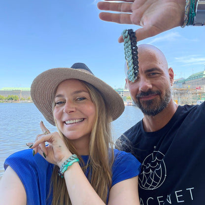 Eine Person trägt das SeaChain Bracenet in 3D Druck aus alten Fischernetzen hergestellt in Blassgrün am Handgelenk und zeigt auf die Alster. Die Person daneben hält zwei weitere SeaChain Bracenets in Blassgrün und Schwarz am Daumen. 