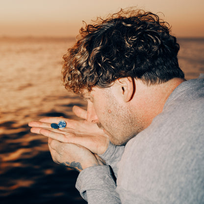 Malte Zierden steht mit zwei "Alles für die Tiere" Ansteck-Pins aus Emaille in Blau und Gold mit "Alles für die Tiere, immer" Schriftaufzug, sowie Robben-Motiv, am Meer.