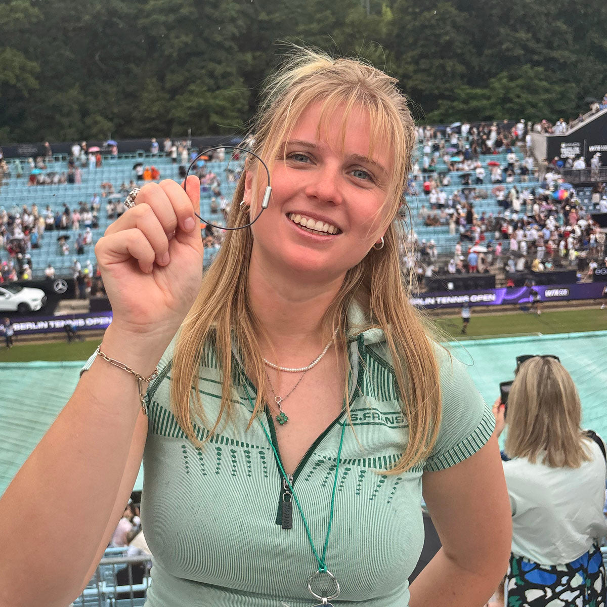 Person hält das CourtNet Armband aus Tennissaiten in Schwarz mit Drehverschluss in Silber vor sich und steht an einem Tennisplatz. 