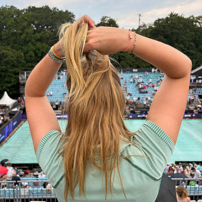 Person steht am Tennisplatz und trägt CourtNet Armband aus Tennissaiten in Pink mit Drehverschluss in Roségold am Handgelenk. Die Person fasst sich in die Haare
