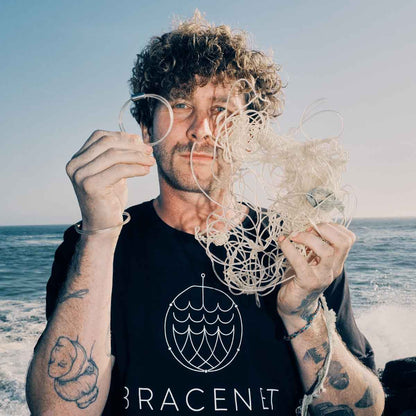 Malte Zierden steht vor dem Meer. In einer Hand hält er das Alles für die Tiere immer - Robbe "Walvis" Armband aus transparenten Langleinen von BRACENET mit Drehverschluss in silber und "Alles für die Tiere, immer" Gravur mit Robben-Motiv. In der anderen Hand hält er die unverarbeiteten Langleinen.