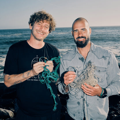 Malte Zierden und Benjamin von BRACENET stehen vor dem Meer. Malte Zierden hält ein grünes Bracenet und grünes Netz in der Hand. Benjamin die transparenten Langeleinen und das Alles für die Tiere immer - Robbe "Walvis" Armband aus transparenten Langleinen von BRACENET mit Drehverschluss in silber und "Alles für die Tiere, immer" Gravur mit Robben-Motiv.