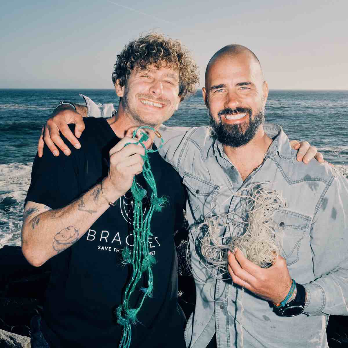 Malte Zierden hält das Alles für die Tiere immer - Robbe Emmanuel Armband aus grün-gelben Fischernetzen von BRACENET mit Bajonettverschluss in roségold in einer Hand und steht Arm in Arm mit Benjamin von BRACENET, der ebenfall Fischernetze in den Armen hält.