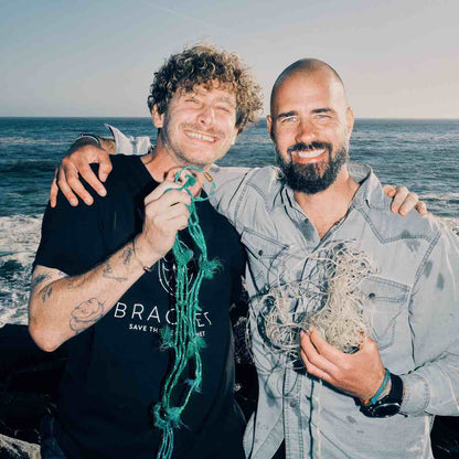 Malte Zierden hält das Alles für die Tiere immer - Robbe Emmanuel Armband aus grün-gelben Fischernetzen von BRACENET mit Bajonettverschluss in roségold in einer Hand und steht Arm in Arm mit Benjamin von BRACENET, der ebenfall Fischernetze in den Armen hält.