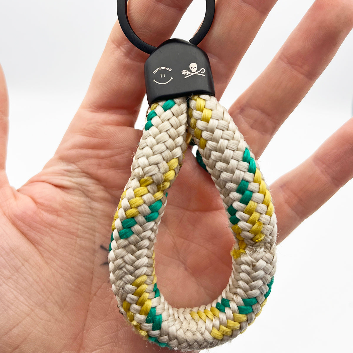 Person hält den Sea Shepherd X BRACENET x humanvoll Schlüsselanhänger mit schwarzer Kappe mit dem Sea Shepherd und humanvoll Logo aus weißem Fischereiseil mit gelben und grünen Akzenten in der Hand