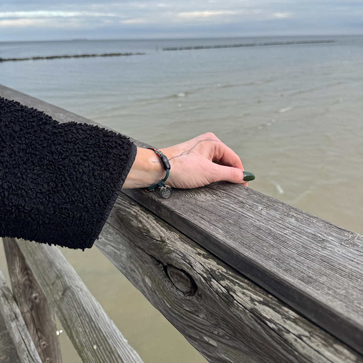 Person trägt das BRACENET Upcycling Armband Insel Rügen aus grünen und bunten Fischernetzen mit schwarzem Verschluss und Anhänger mit Inselgravur am Handgelenk und hat den Arm auf dem Geländer der Seebrücke liegen. 