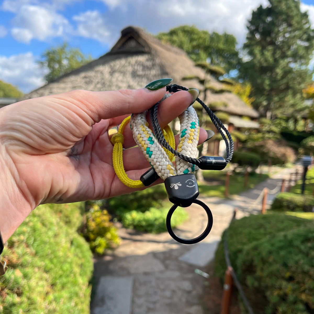 Person hält Sea Shepherd X BRACENET x humanvoll Armband aus schwarzen Fischernetzen mit schwarzem Magnetverschluss auf dem das Sea Shepherd Logo, das humanvoll Logo und das BRACENET Logo graviert sind zusammen mit den anderen Produkten dieser kollektion in der Hand. Im Hintergrund sieht man ein Haus
