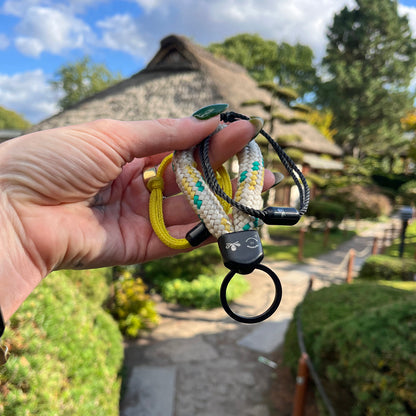 Person hält Sea Shepherd X BRACENET x humanvoll Armband aus schwarzen Fischernetzen mit schwarzem Magnetverschluss auf dem das Sea Shepherd Logo, das humanvoll Logo und das BRACENET Logo graviert sind zusammen mit den anderen Produkten dieser kollektion in der Hand. Im Hintergrund sieht man ein Haus