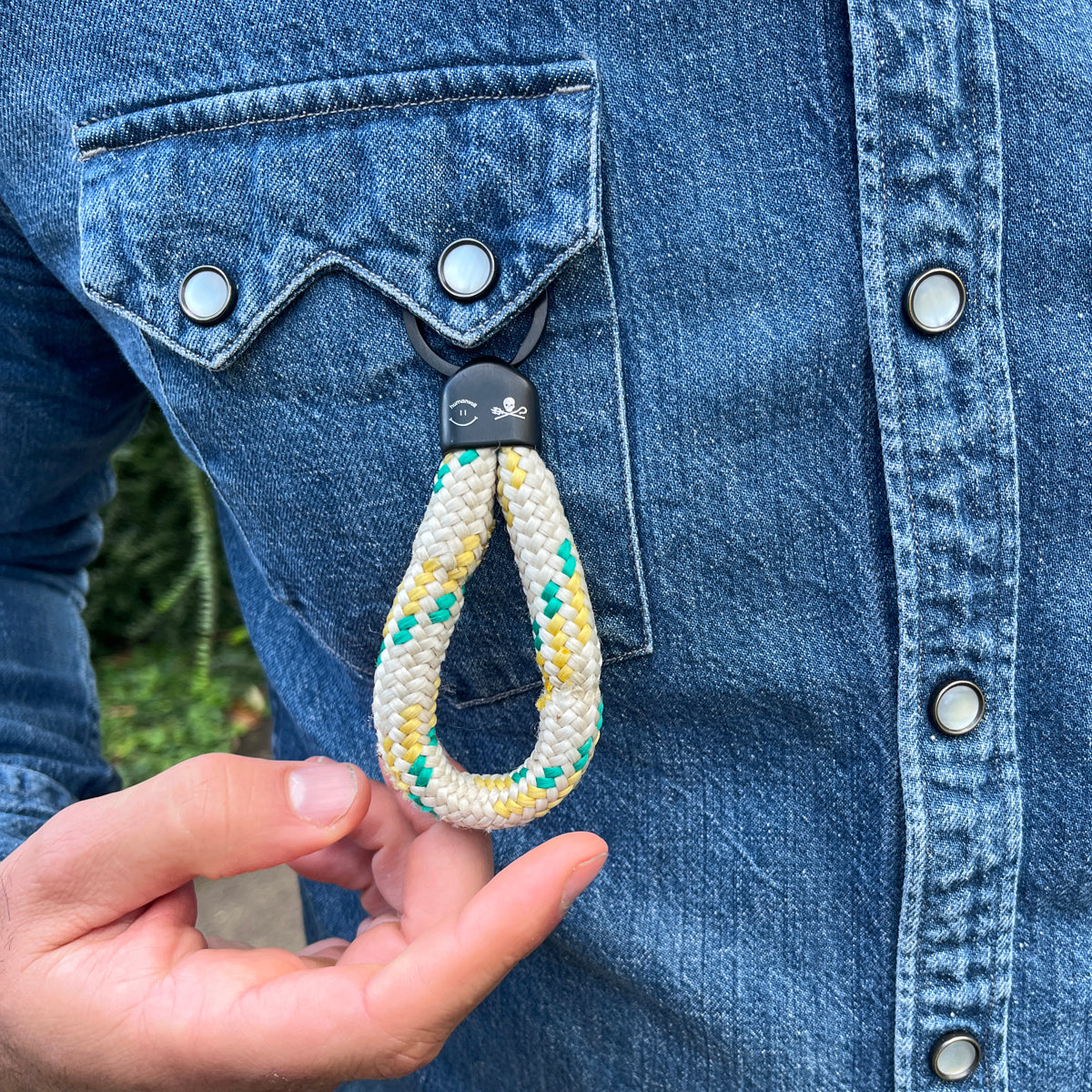 Der Sea Shepherd X BRACENET x humanvoll Schlüsselanhänger mit schwarzer Kappe mit dem Sea Shepherd und humanvoll Logo aus weißem Fischereiseil mit gelben und grünen Akzenten hängt an der Brusttasche von einem Hemd