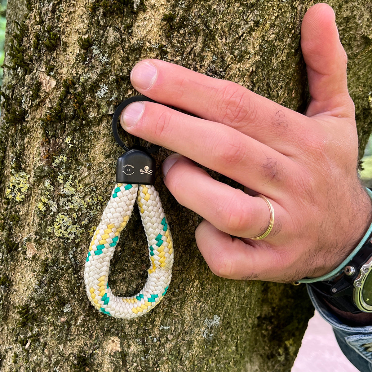 Person hält den Sea Shepherd X BRACENET x humanvoll Schlüsselanhänger mit schwarzer Kappe mit dem Sea Shepherd und humanvoll Logo aus weißem Fischereiseil mit gelben und grünen Akzenten an einen Baum